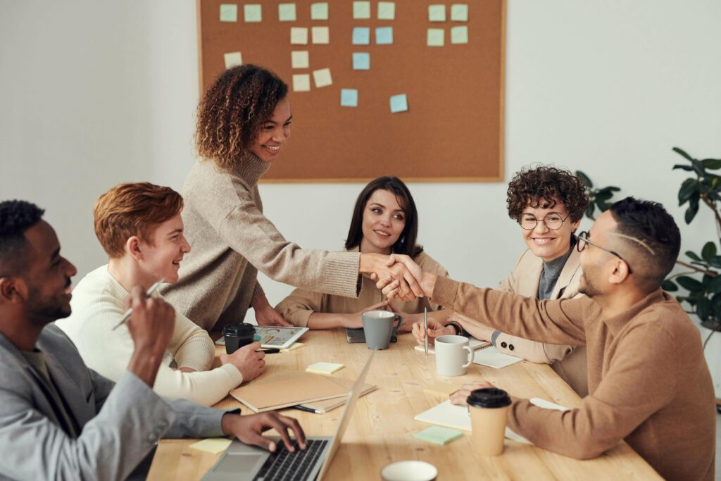 A group of young professionals are sat around a long wooden office desk. They are all smiling while 2 of the 6 individuals shake hands. There is a laptop, books and coffee cups across the desk.