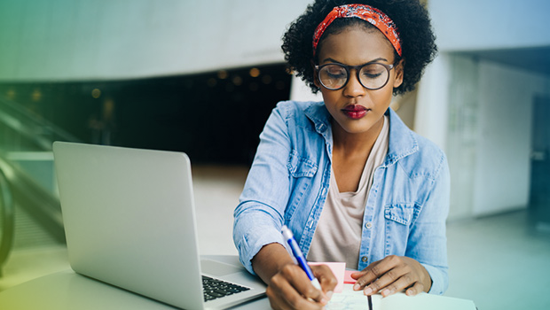 A woman is sat at a desk with her laptop open. She is writing at a notebook with a pen. She is wearing large thick framed glassed and a denim shirt.