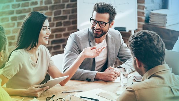 A team of 6 sit around a table engaged in conversation. The man at the head of the able has his laptop open and one of the women at the table has a table. She is smiling as everyones eyes focus on her.