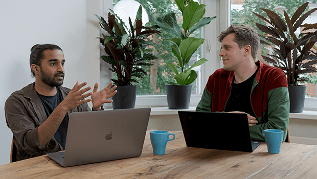 Two men are sat in an office space with their laptops in front of them. They are sat at a wooden desk and are surrounded by plants.
