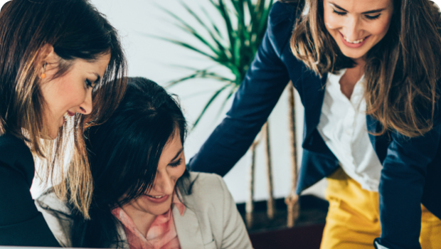 A women is standing over two women in an office setting. They are all smiling and looking over a document.