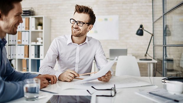 A man is holding a document and point to it with a pencil while smiling. He is showing the document to another man. Both men are dressed in smart casual attire and sat in a modern office.