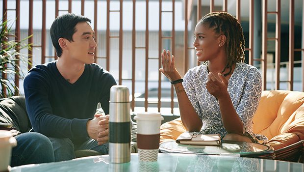 A man and a woman are sat on soft chairs engaged in conversation. There are reusable coffee cups on the desk in front of them.